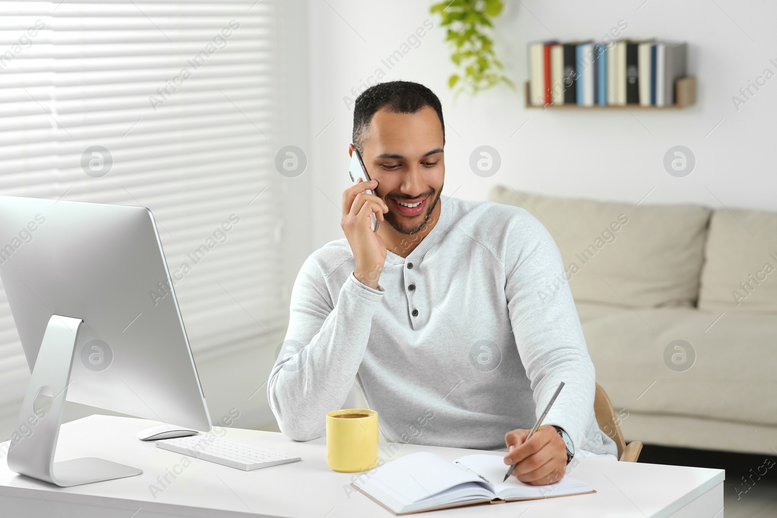 Photo of Young man talking on smartphone while working with computer at desk in room. Home office