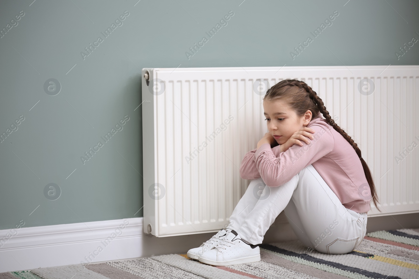 Photo of Sad little girl suffering from cold on floor near radiator