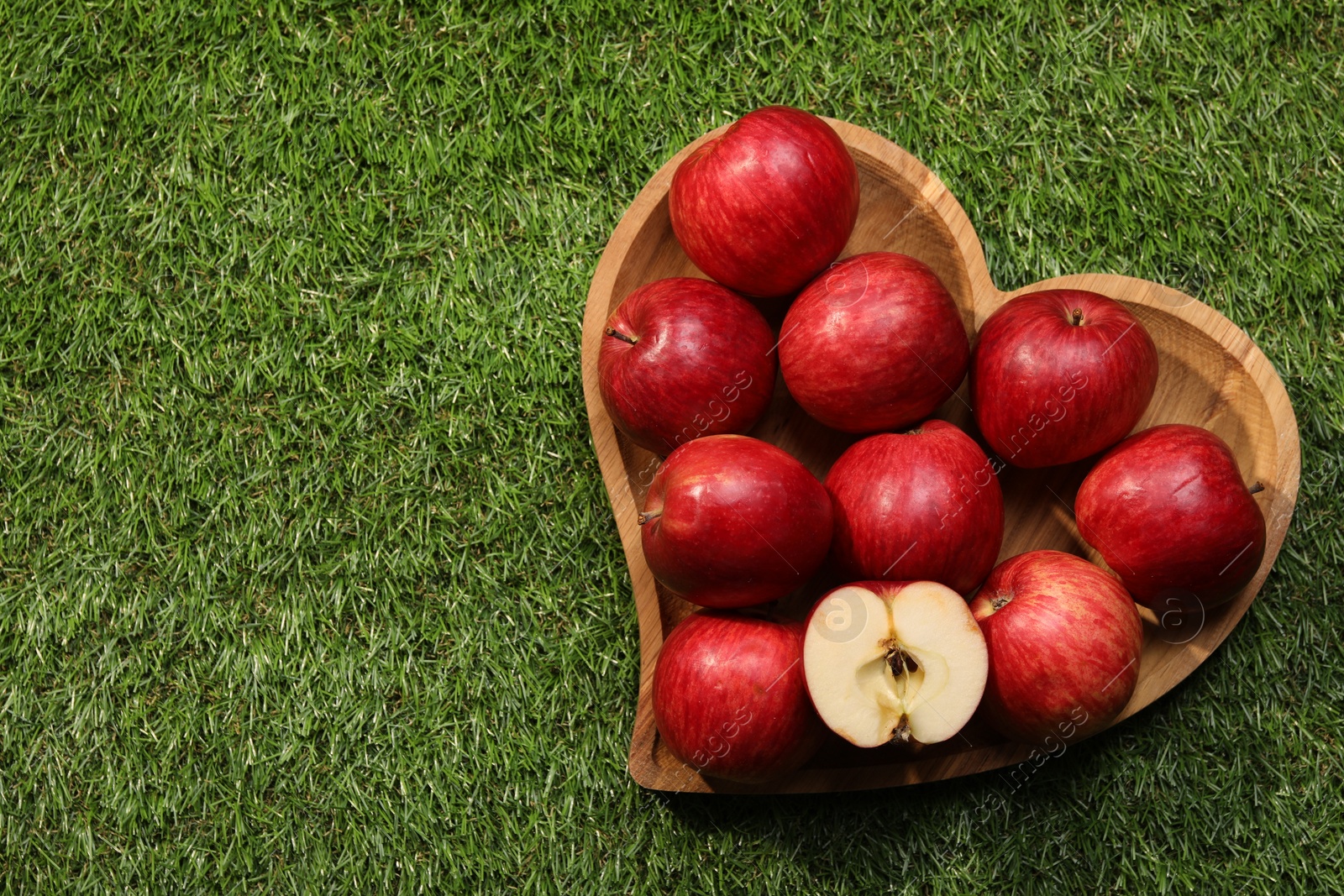 Photo of Wooden plate in shape of heart with red apples on green grass, top view. Space for text