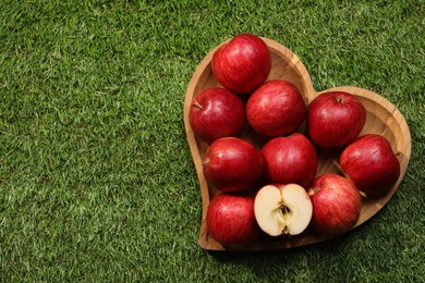 Photo of Wooden plate in shape of heart with red apples on green grass, top view. Space for text