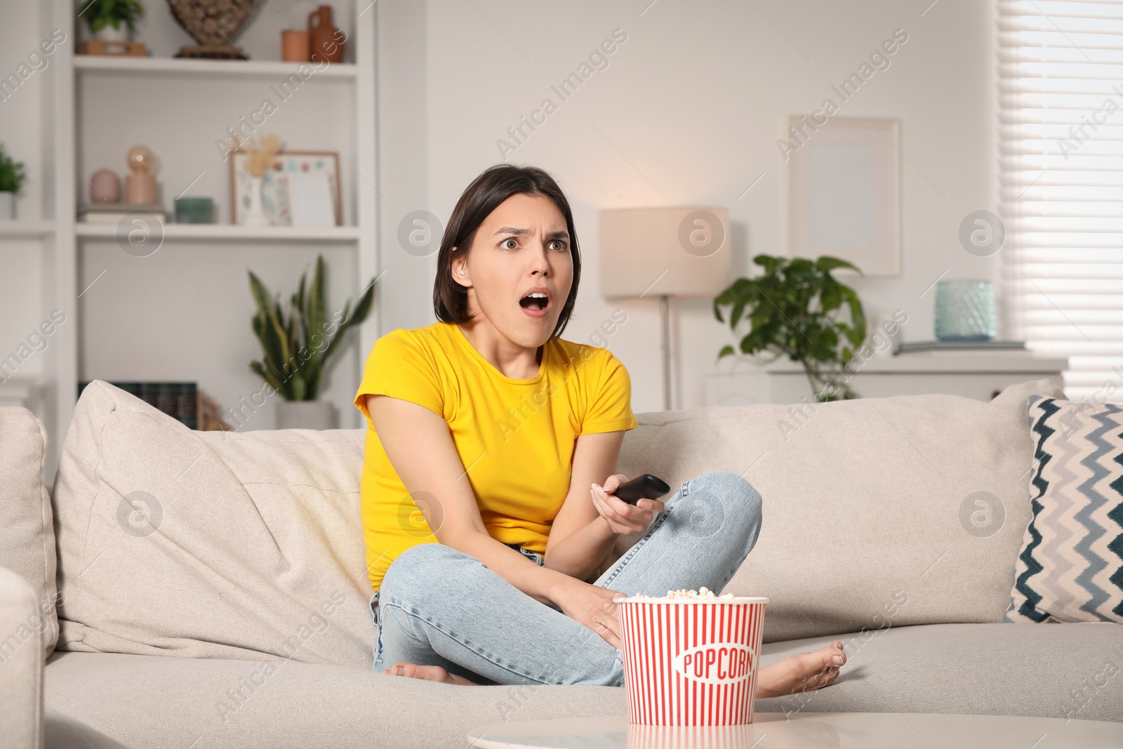 Photo of Surprised woman watching TV with popcorn on sofa at home