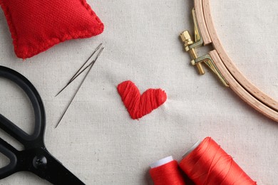 Photo of Embroidered red heart, needles, hoop and scissors on light cloth, top view