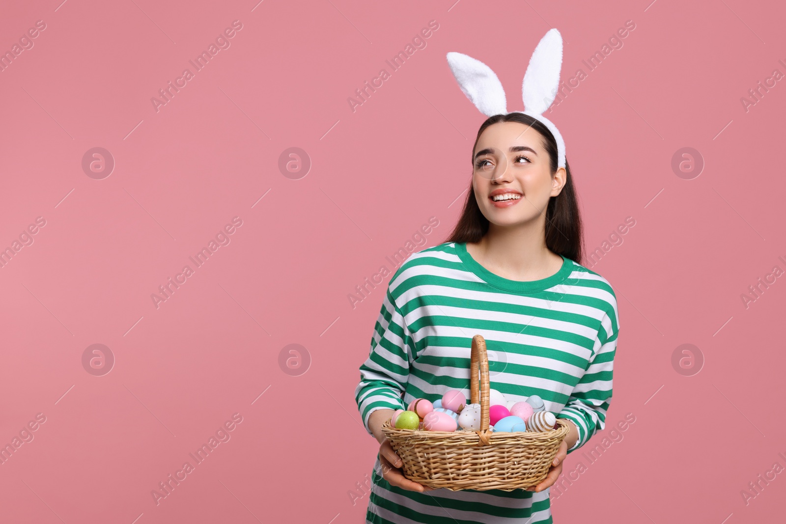 Photo of Happy woman in bunny ears headband holding wicker basket of painted Easter eggs on pink background. Space for text