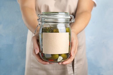 Photo of Woman holding jar of pickled cucumbers with blank sticker against blue background, closeup view. Space for text
