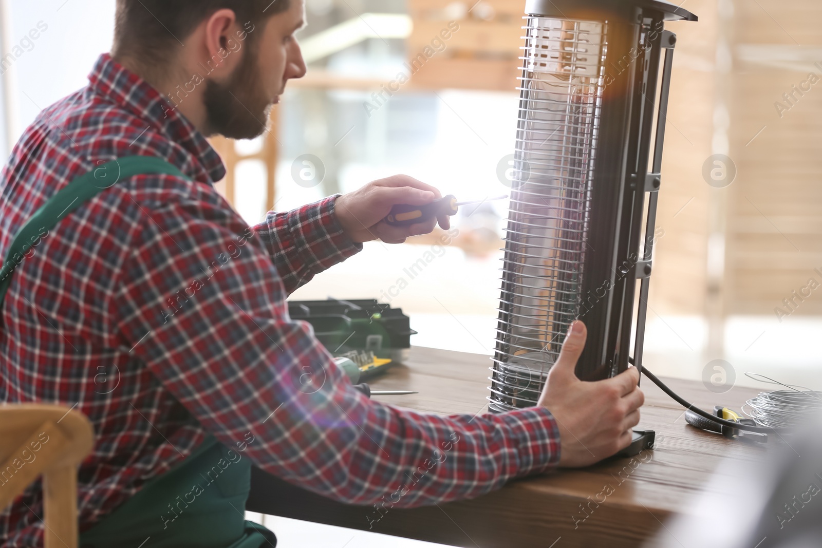 Photo of Professional technician repairing electric halogen heater with screwdriver at table indoors