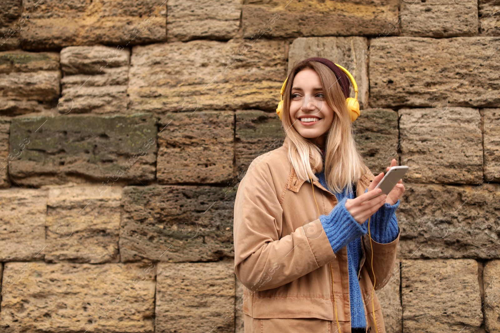 Photo of Young woman with headphones listening to music near stone wall. Space for text