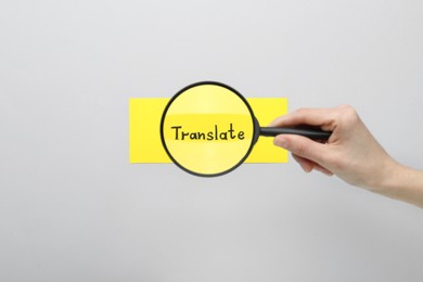 Photo of Woman holding magnifying glass over sheet of paper with word Translate on light grey background, top view
