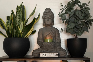 Photo of Buddha sculpture with candle and green houseplants on shelf indoors. Interior design