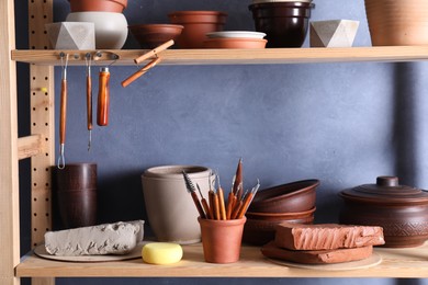 Photo of Set of different crafting tools and clay dishes on wooden rack in workshop