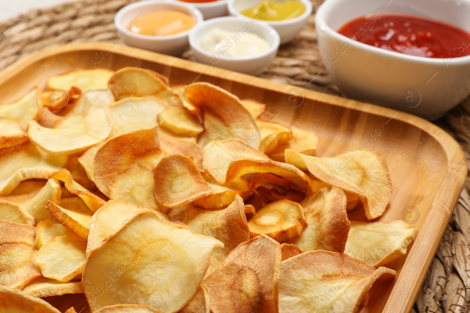 Photo of Tasty homemade parsnip chips with different sauces on wicker surface, closeup