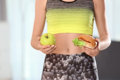 Photo of Woman holding tasty sandwich and fresh apple, indoors. Choice between diet and unhealthy food