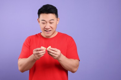 Photo of Happy asian man holding tasty fortune cookie with prediction on purple background. Space for text