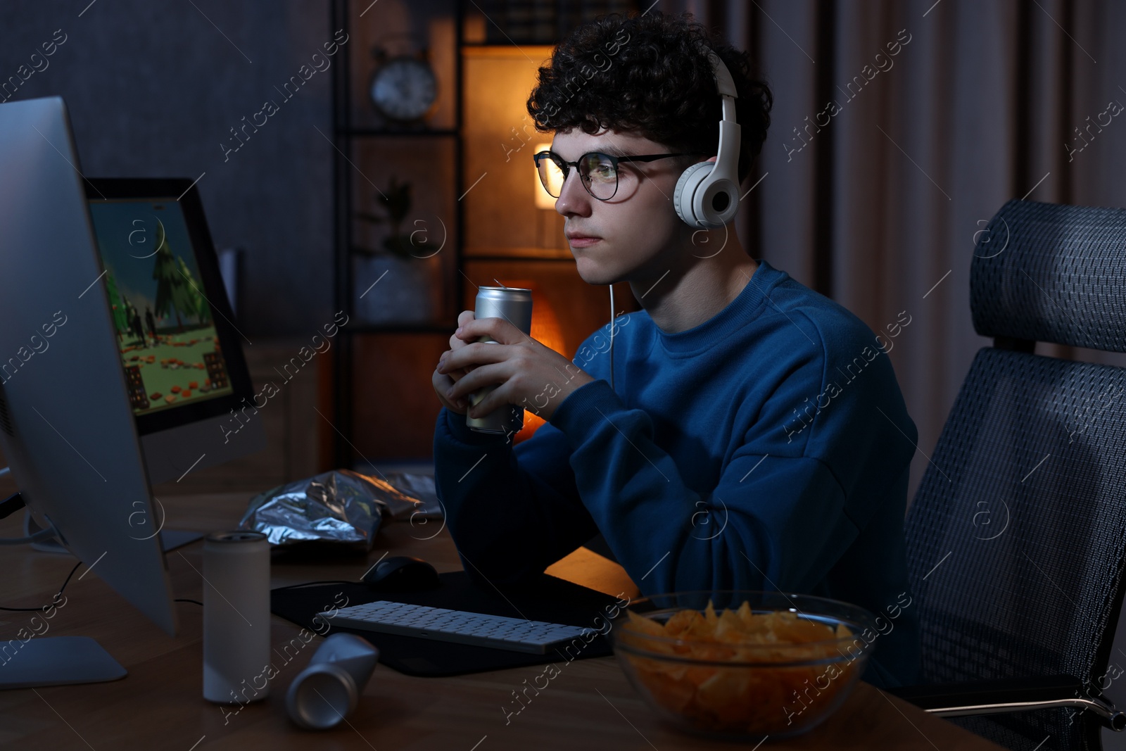 Photo of Young man with energy drink and headphones playing video game at wooden desk indoors