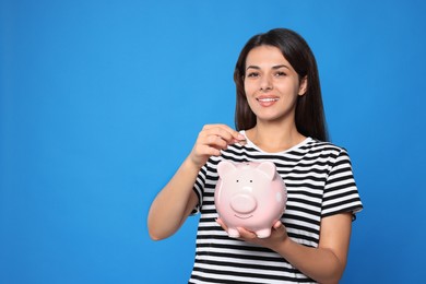 Young woman putting coin into piggy bank on light blue background, space for text