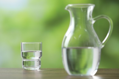 Jug and glass with clear water on wooden table against blurred green background, selective focus