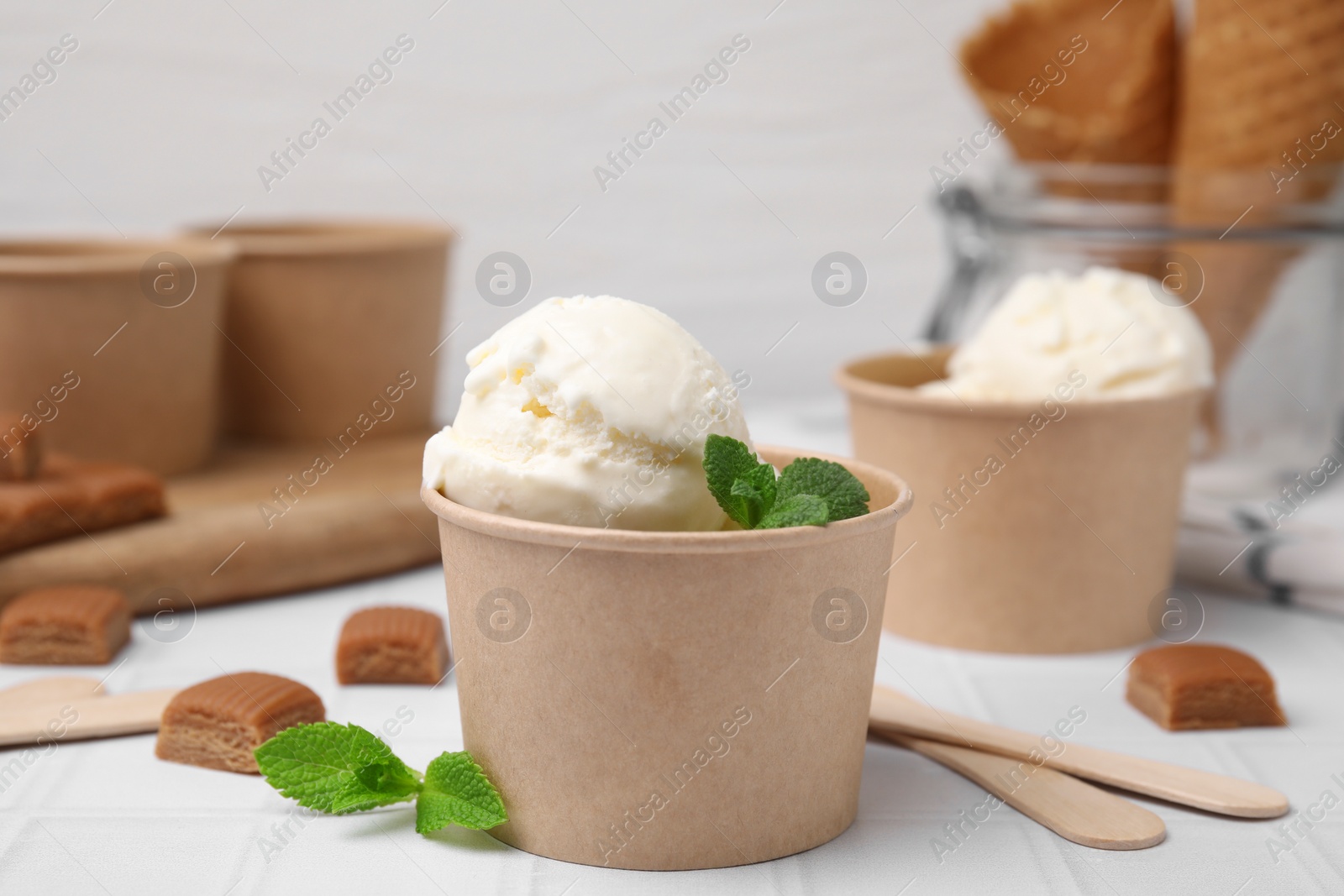 Photo of Scoops of ice cream with caramel sauce, mint leaves and candies on white tiled table, closeup