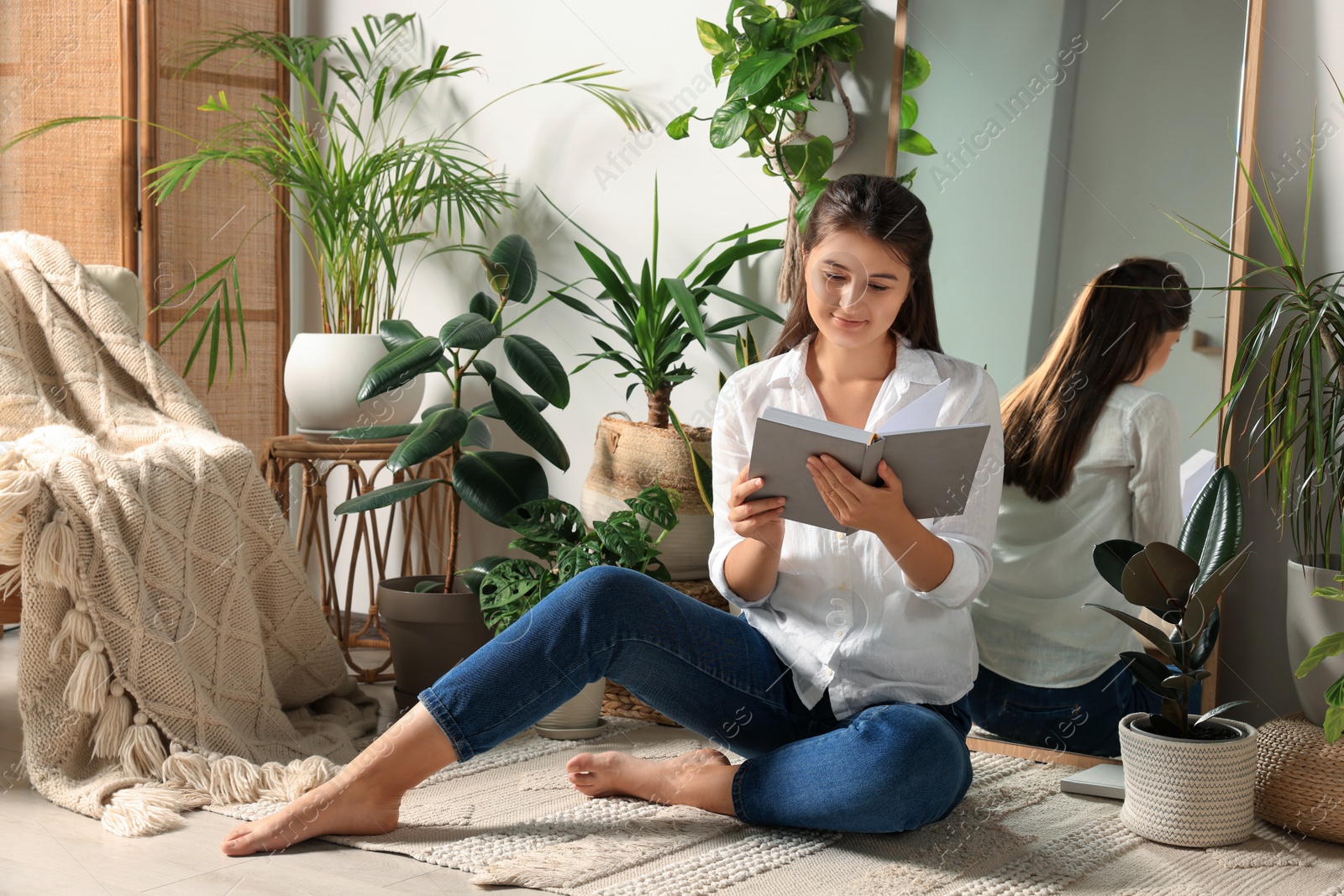 Photo of Happy young woman reading book near mirror and different houseplants on floor in room