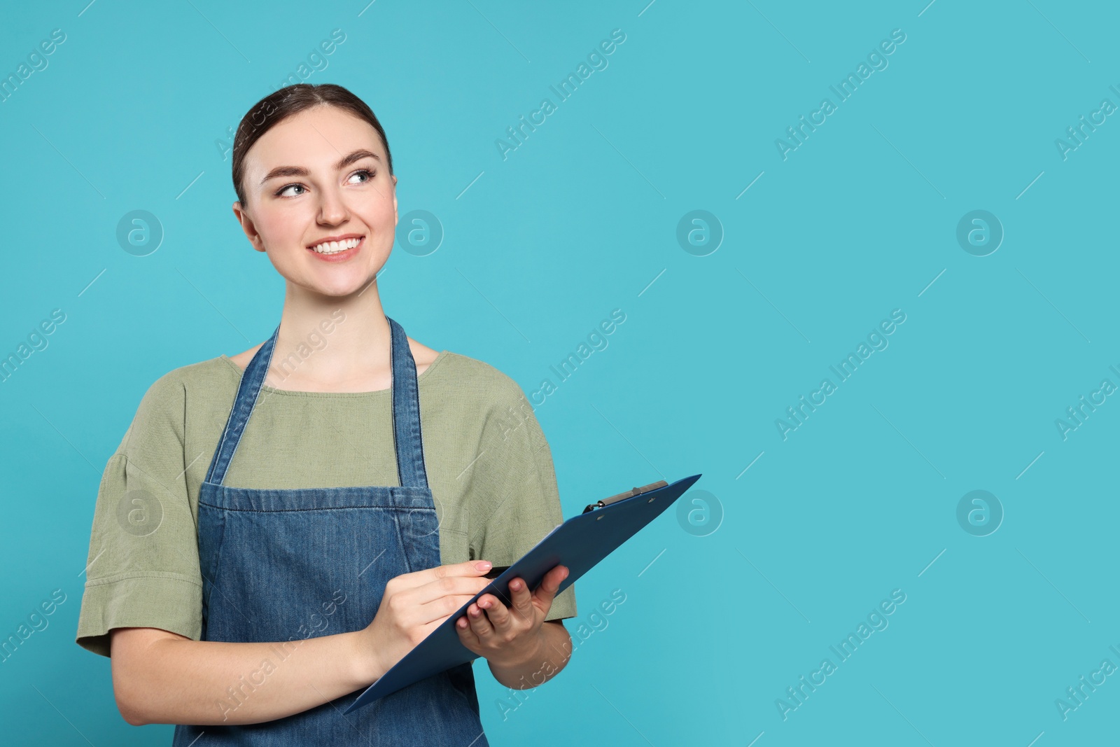 Photo of Beautiful young woman in clean denim apron with clipboard on light blue background. Space for text