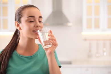 Young woman drinking pure water from glass in kitchen. Space for text