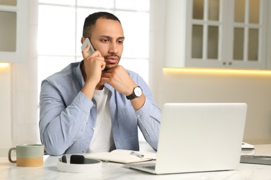 Photo of Young man talking on smartphone while working with laptop at desk in kitchen. Home office