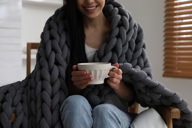 Photo of Woman with chunky knit blanket and cup in armchair at home, closeup