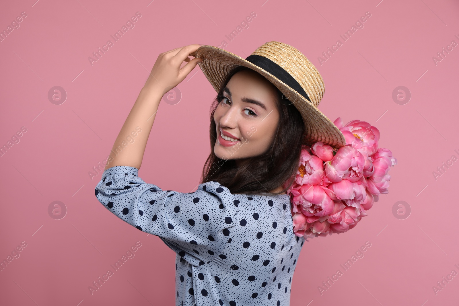 Photo of Beautiful young woman in straw hat with bouquet of peonies on pink background