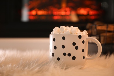 Photo of Cup of hot drink with marshmallows on fluffy rug near fireplace at home. Space for text