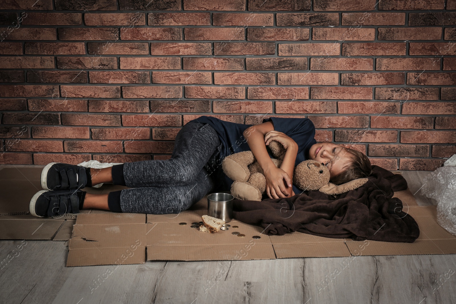 Photo of Poor homeless boy sleeping with his toy on floor near brick wall