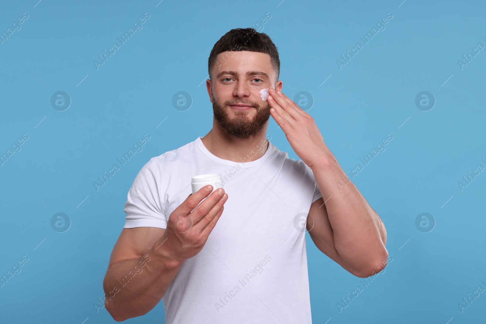 Photo of Handsome man applying cream onto his face on light blue background