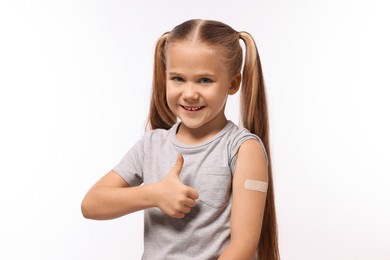 Photo of Happy girl with sticking plaster on arm after vaccination showing thumbs up against white background