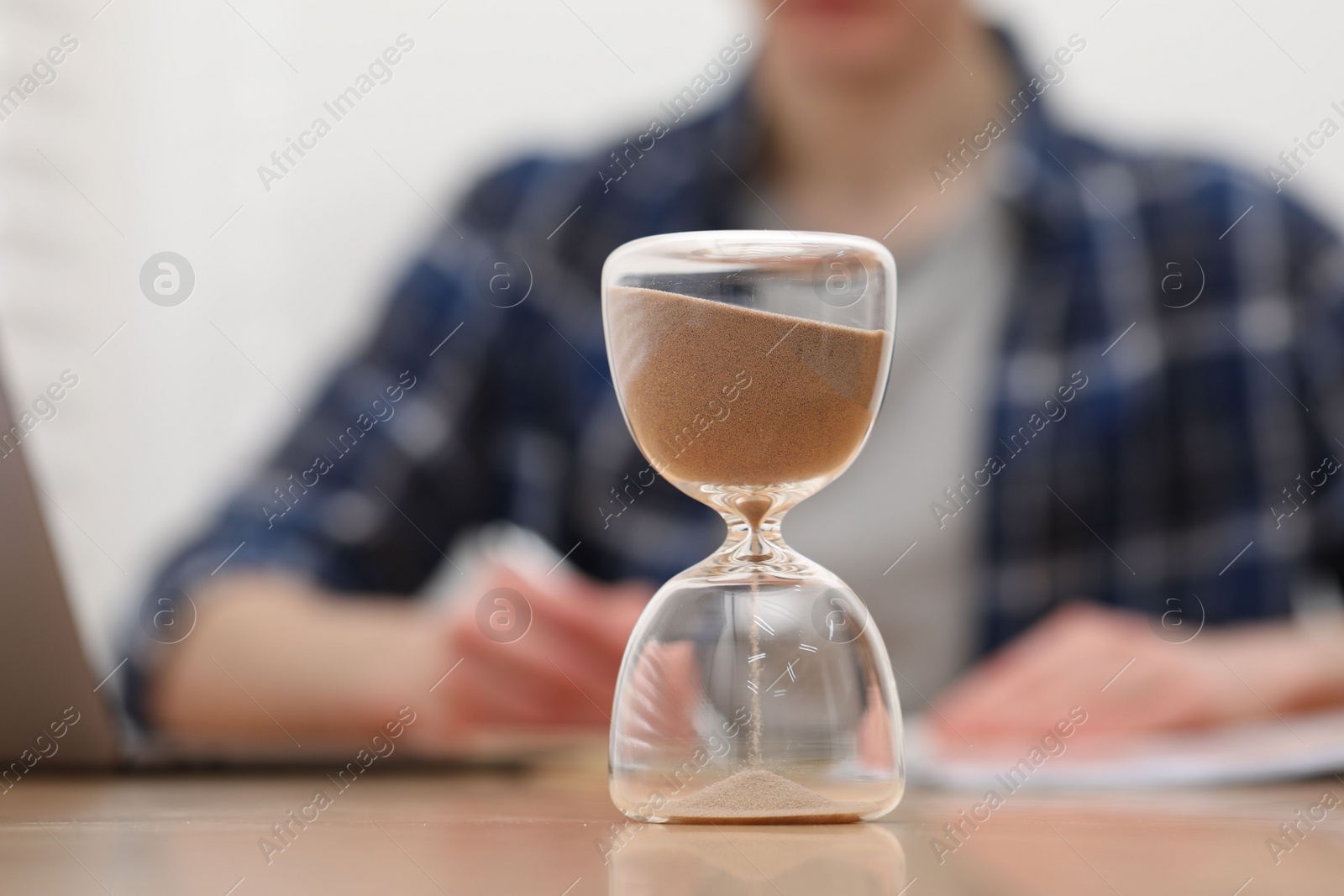 Photo of Hourglass with flowing sand on desk. Man taking notes indoors, selective focus