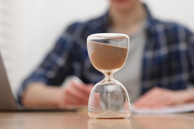 Photo of Hourglass with flowing sand on desk. Man taking notes indoors, selective focus