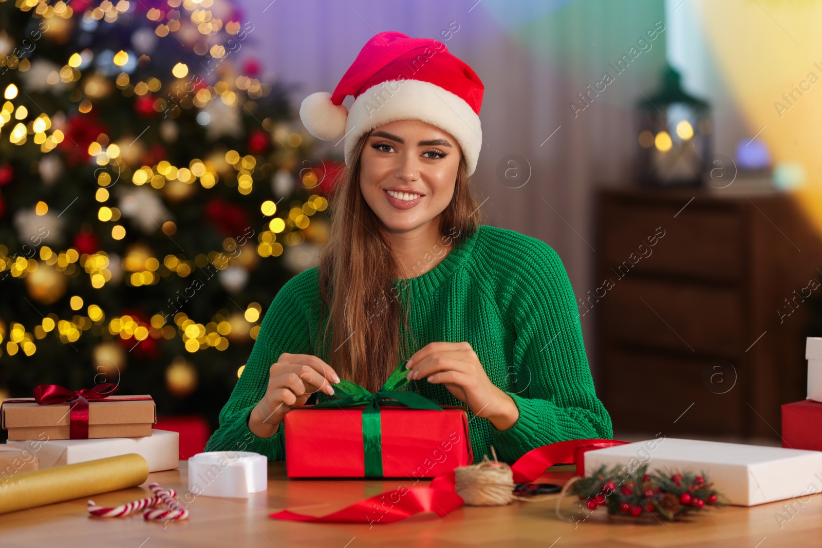 Photo of Beautiful young woman in Santa hat making Christmas gift at table in room