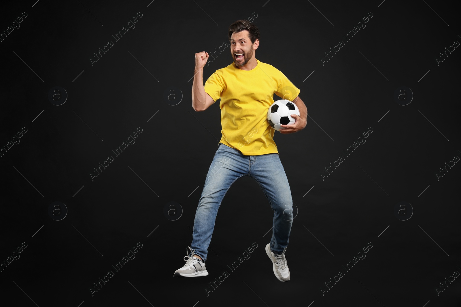 Photo of Emotional sports fan with soccer ball jumping on black background