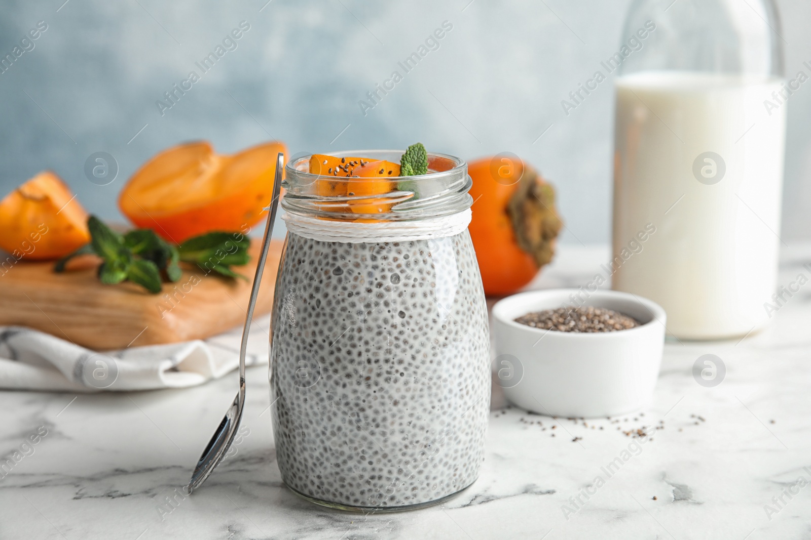 Photo of Glass jar of tasty chia seed pudding with persimmon and ingredients on table