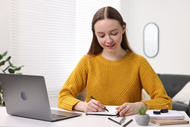 Photo of E-learning. Young woman taking notes during online lesson at white table indoors