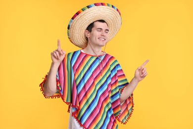 Photo of Young man in Mexican sombrero hat and poncho pointing at something on yellow background