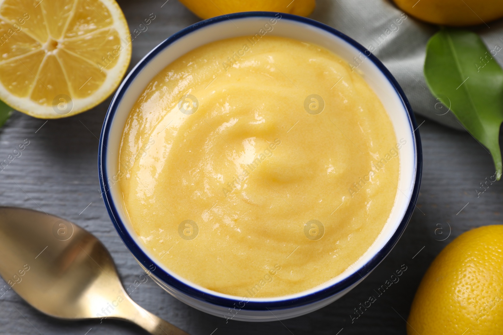 Photo of Delicious lemon curd in bowl, fresh citrus fruits, green leaf and spoon on grey wooden table, closeup