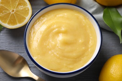 Delicious lemon curd in bowl, fresh citrus fruits, green leaf and spoon on grey wooden table, closeup