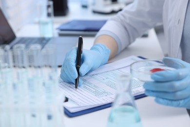 Photo of Laboratory worker holding petri dish with blood sample while working at white table, closeup