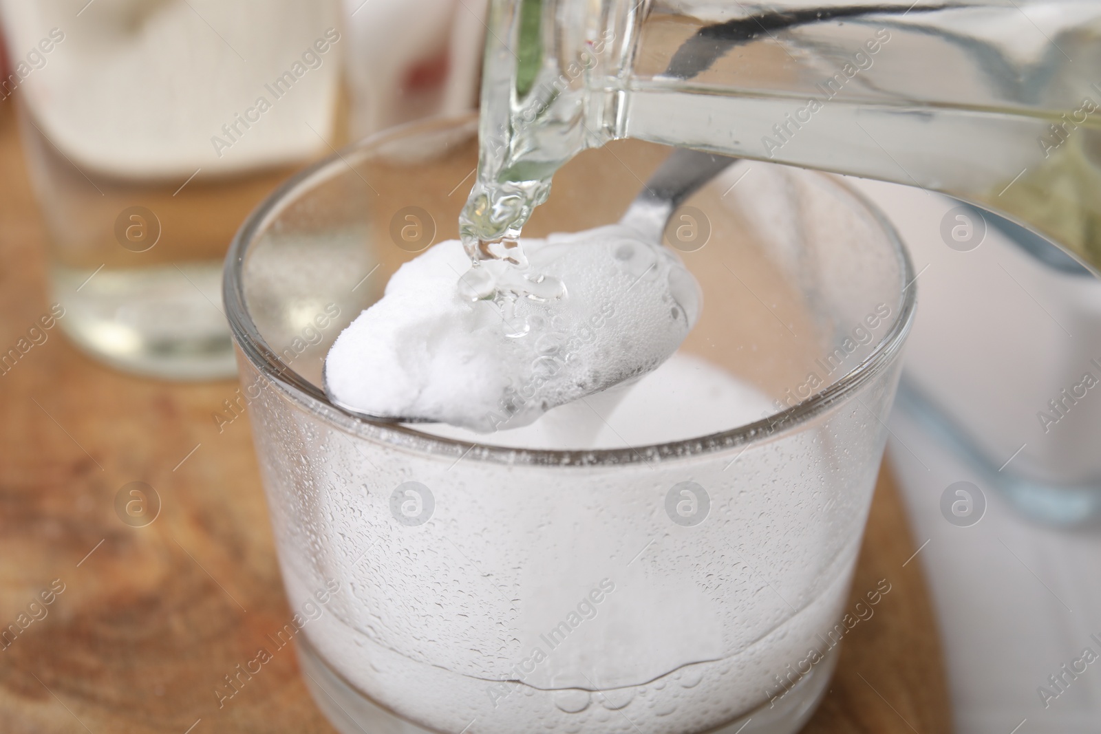 Photo of Pouring vinegar into spoon with baking soda over glass bowl at table, closeup