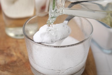 Photo of Pouring vinegar into spoon with baking soda over glass bowl at table, closeup