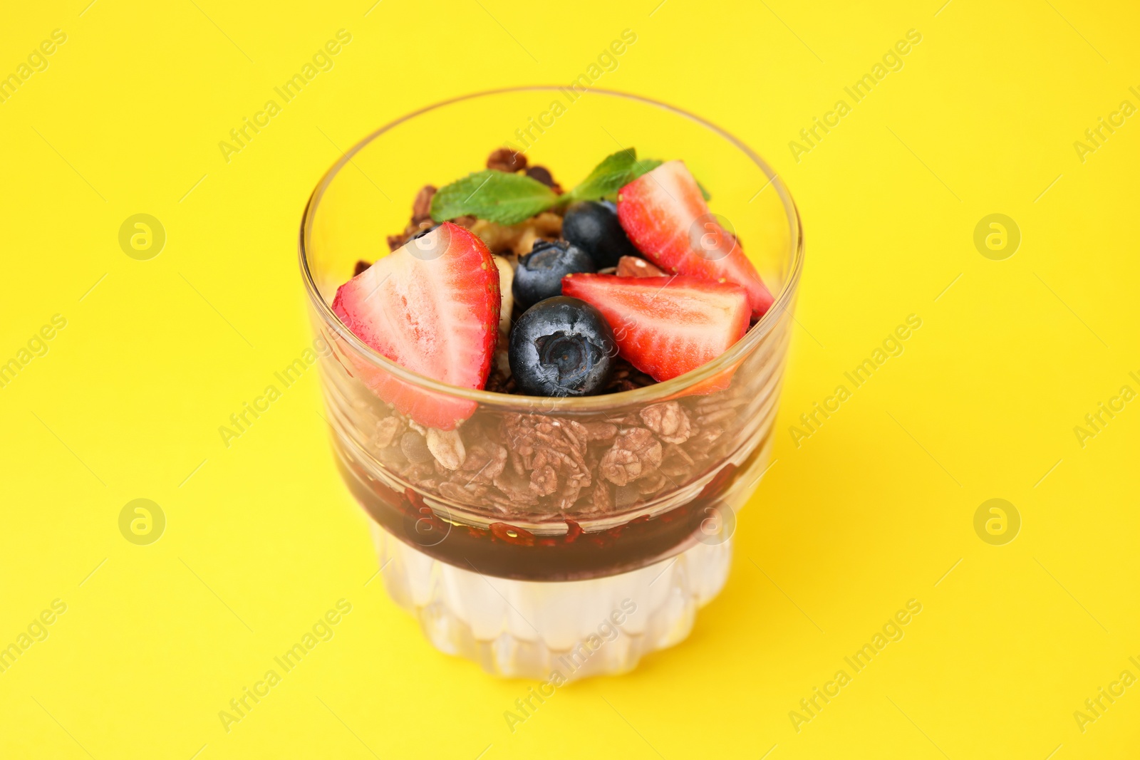 Photo of Tasty granola with berries, jam and yogurt in glass on yellow background, closeup