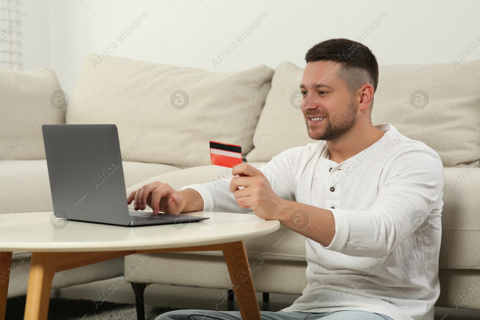 Photo of Happy man with credit card using laptop for online shopping at white table in room