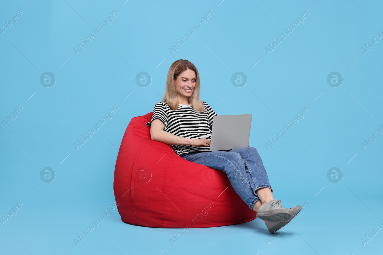 Photo of Happy woman with laptop sitting on beanbag chair against light blue background