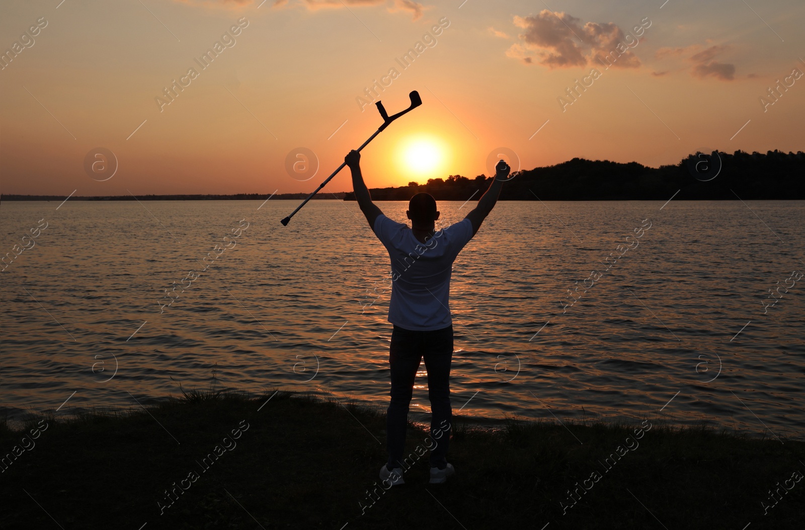 Photo of Man raising elbow crutch up to sky near river at sunset, back view. Healing miracle