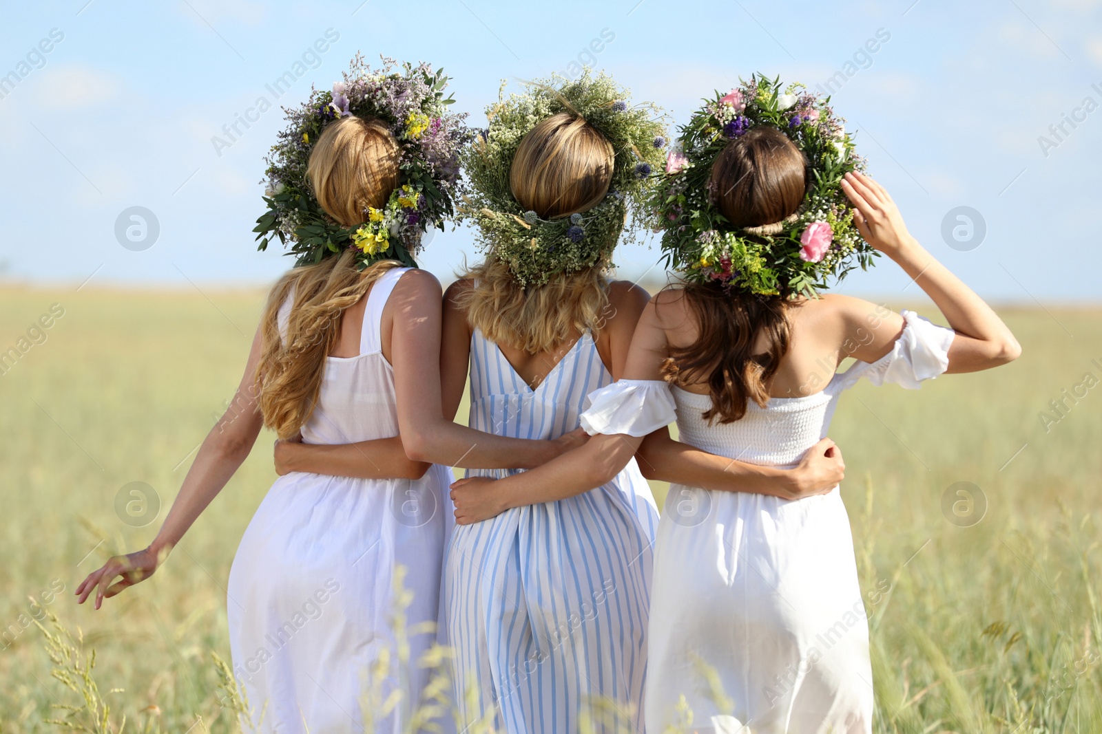 Photo of Young women wearing wreaths made of beautiful flowers in field on sunny day, back view