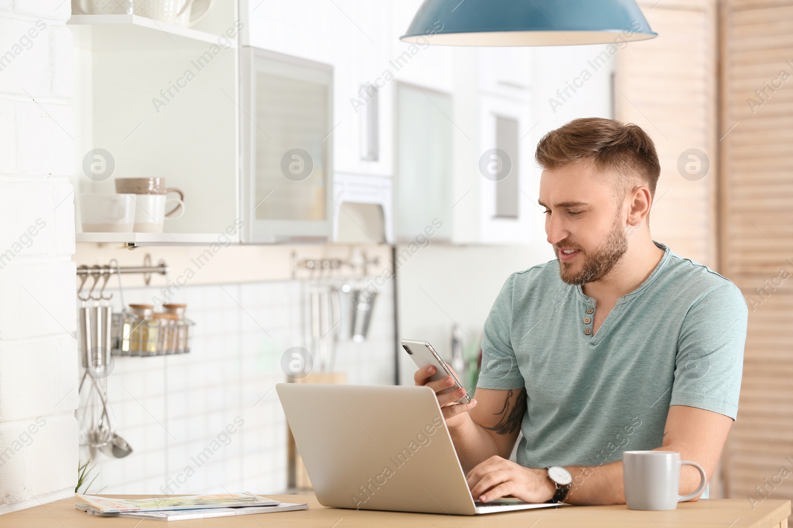 Photo of Young man using mobile phone and laptop at table in kitchen