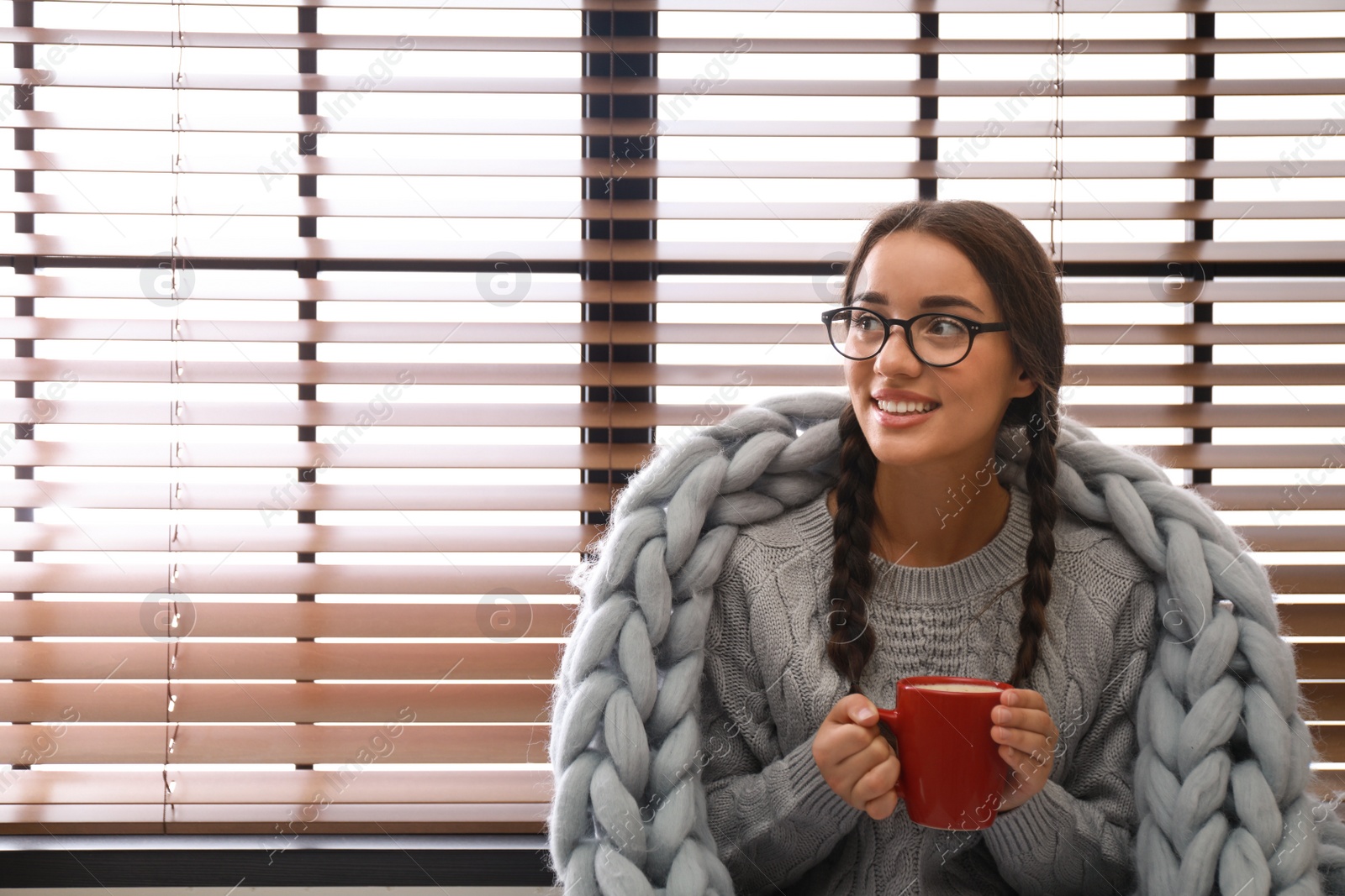 Image of Happy young woman wrapped in knitted blanket with cup of hot drink near window. Lazy morning