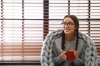 Happy young woman wrapped in knitted blanket with cup of hot drink near window. Lazy morning